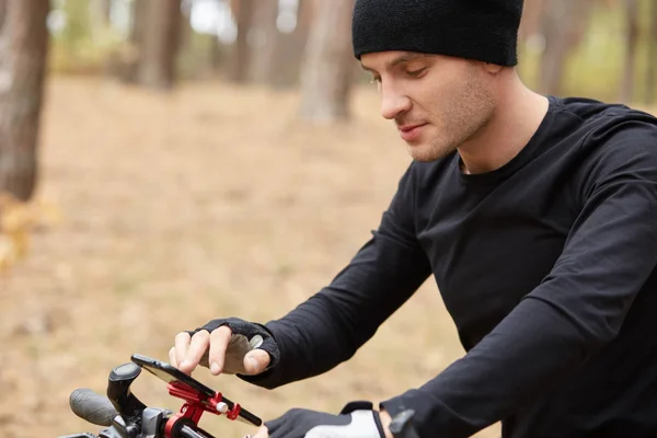 Közelről kültéri ortrait a fiatal férfi kerékpáros segítségével mobiltelefon utcán, megtalálás útját az erdőben mobiltelefonon keresztül, vonzó ember visel fekete kerékpáros ruhákat, kiadási idő lovaglás Bike. — Stock Fotó
