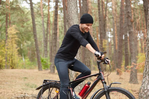 Venkovní záběr mladého pohledného muže, který jede na kole na přírodní pozadí, je šťastný a dívá se na své zařízení připevněné k bicyklu, muži nosí černé oblečení. Koncepce zdravého životního stylu. — Stock fotografie