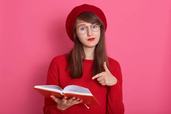 Foto horizontal de mujer con libreta naranja apuntando a sus páginas con su dedo índice. Atractiva hembra con suéter rojo, boina y gafas, que tiene la piel perfecta y maquillaje de granada brillante . — Foto de Stock