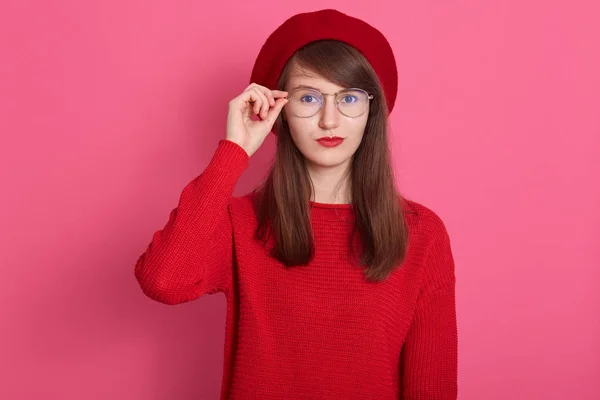 Imagem de estudantes do sexo feminino bonita vestindo roupas vermelhas e boina, tem expressão facial séria, olhando atentamente para a câmera, mantém a mão em seus óculos, posando isolado sobre fundo rosa . — Fotografia de Stock