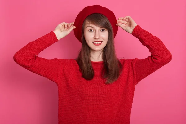 A mulher bonita na moda no estilo francês mantém as mãos em sua cobertura para a cabeça, olha para a câmera, tendo bom humor, vestindo roupas vermelhas e boina, posando com expressão facial agradável. Conceito de pessoas . — Fotografia de Stock