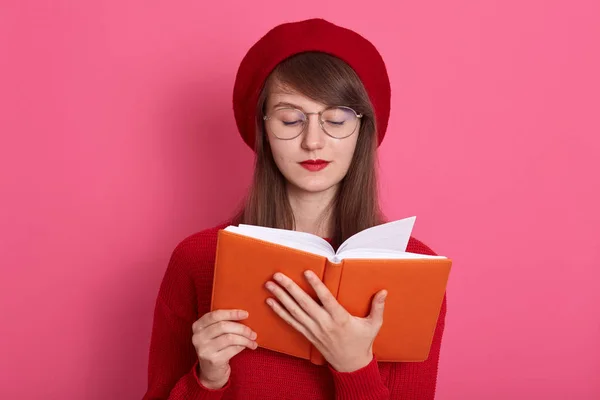 Imagen de una humilde morena vestida con boina roja, suéter y gafas, libro de lectura aislado sobre fondo rosado en el estudio, mirando a la cámara con expresión facial concentrada. Concepto educativo . —  Fotos de Stock