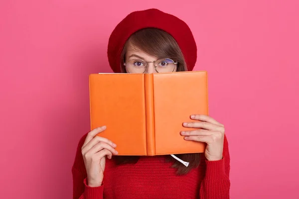 De cerca retrato de mujer joven esconde su cara detrás de su cuaderno portátil naranja, encantadora mujer lleva suéter rojo, boina y gafas, se ve asustada detrás de su organizador, aislado en la pared rosa . — Foto de Stock