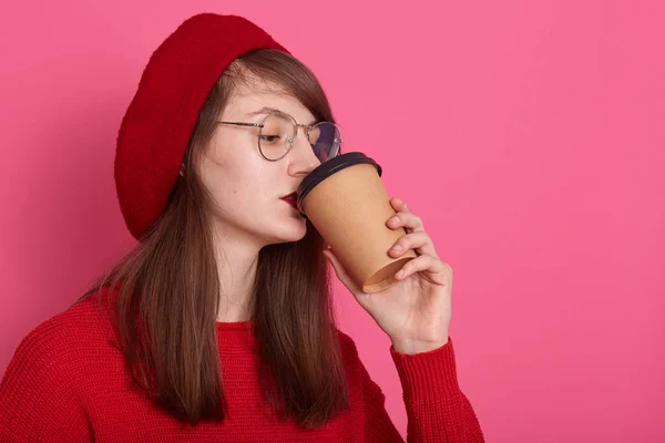 Estudio de tiro de mujer hermosa viste camisa roja, gafas y boina, sosteniendo taza de café en taza de plástico, perfil de chica encantadora bebiendo baño caliente. Copiar espacio para publicidad o promoción . — Foto de Stock