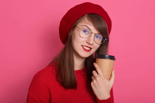 Εσωτερική έλλειψη ελκυστική γυναίκα σε κόκκινο μπερέ, πουλόβερ και γυαλιά, κρατώντας φλιτζάνι καφέ ή τσάι σε χέρια που απομονώνονται σε ροζ φόντο στούντιο, κοιτάζοντας χαμογελώντας απευθείας στην κάμερα. Έννοια ανθρώπων. — Φωτογραφία Αρχείου