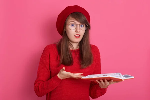Retrato de una hermosa chica vestida con jersey rojo y suéter, sosteniendo libros, mira a la cámara con los ojos y la boca ampliamente abiertos, modelo posando aislado sobre fondo rosa. Concepto de personas y educación . — Foto de Stock