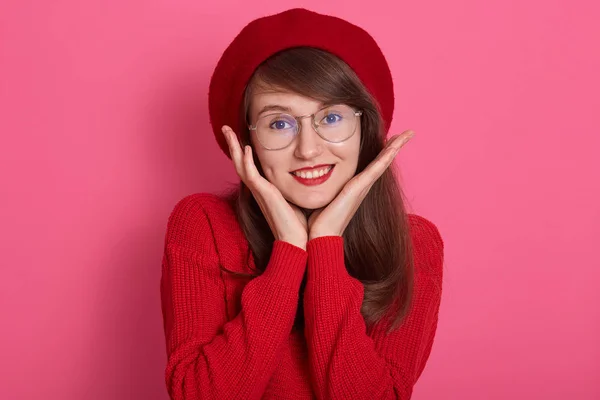 Primer plano retrato de una bella mujer sonriente, expresa felicidad, adorable chica con gafas redondeadas, tocándose las mejillas con las palmas de las manos, llevando boina roja y suéter, mirando directamente a la cámara . — Foto de Stock