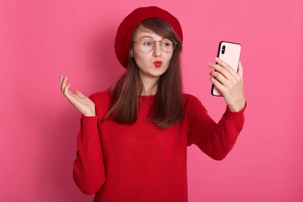 Foto dalam ruangan dari wanita Kaukasia yang mempesona membuat gerakan ciuman sambil memotret dirinya sendiri. Potret romantis wanita berambut gelap gaun baret dan sweater, membuat selfie atas latar belakang merah muda . — Stok Foto