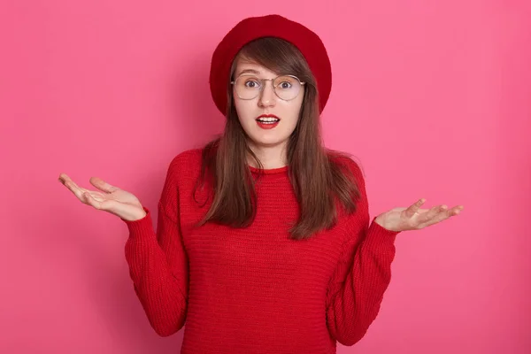 Potret gadis bingung mengenakan baret merah, sweater dan kacamata bulat, menyebarkan tangannya ke samping, keepsmouth terbuka, terisolasi di atas latar belakang dinding merah muda. Orang-orang emosi tulus, konsep gaya hidup — Stok Foto