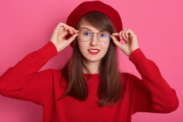 Imagem da mulher bonita na moda em estilo francês mantém o dedo em seus óculos, olha para a câmera, tendo bom humor, vestindo camisola vermelha e boina, mulher está sorrindo contra a parede do estúdio rosa . — Fotografia de Stock