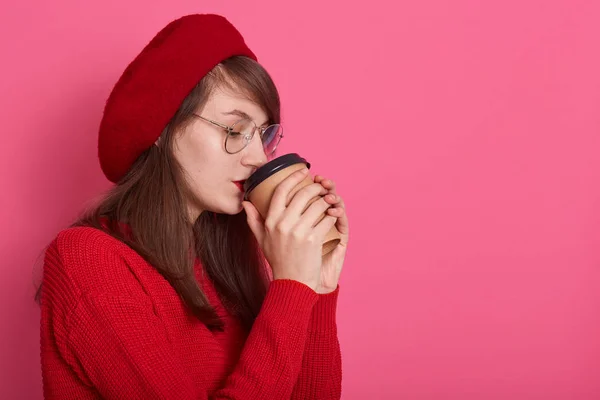 Vista lateral da jovem mulher de cabelos escuros pensativo com copo descartável de café takeaway, de pé contra camisola de camisola rosa fundo dressess e boina vermelha, espaço de cópia para publicidade ou promoção . — Fotografia de Stock