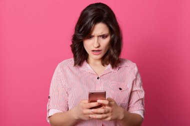 Şok edici mesaj konsepti. Şaşkın, genç, siyah saçlı bir kadın cep telefonu tutuyor ve gül arkaplanının karşısında dikilirken ekranına bakıyor, yüz ifadesini şaşırtıyor. Teknoloji konsepti.