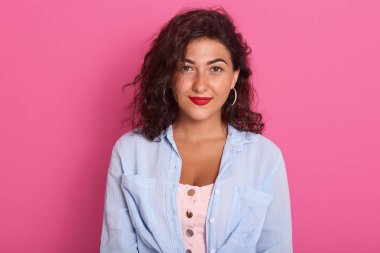 Güzel bir kadının portresi, güzel neşeli bir kız koyu dalgalı saçları ve temiz bir teni var, şık kıyafetler giyiyor, pembe arka planda izole edilmiş bir şekilde poz veriyor, kameraya gülümsüyor..
