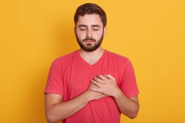 Sakallı erkeğin yatay görüntüsü hoş görünüyor, ellerini kalbinde tutuyor, insanlara karşı iyi hislerini ifade ediyor, sakin bir yüz ifadesi var, sarı stüdyo arka planında izole edilmiş..