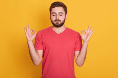 Çekici bir adamın portresi, kırmızı rahat bir tişört giyiyor. Kapalı gözler ve iki elle işaret veriyor. Stüdyoda sarı arka planda izole edilmiş bir model. Meditasyon kavramı.