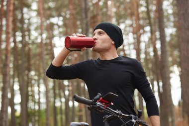 Ormanda dağ bisikleti süren, içme suyu içmek için kısa bir mola veren siyah spor giysili ve şapkalı, ağaçların etrafında poz veren bir adamın yatay görüntüsü. Spor, sağlıklı yaşam tarzı ve doğa konsepti.