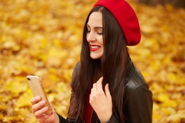Sonbaharda cep telefonuyla selfie çeken bir kadın. Sonbahar kızı akıllı telefon görüntülü konuşma yapıyor. Ormanda sonbahar renklerinde beyaz kadın portresi, kırmızı bereli ve ceketli kadın..