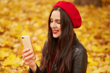 Genç bir kadının selfie çekerken ya da sonbahar parkında görüntülü konuşma yaparken. Güzel kız kırmızı bereli, deri ceketli, akıllı telefonuyla fotoğraf çeken, altın yaprakları örten bir kız.