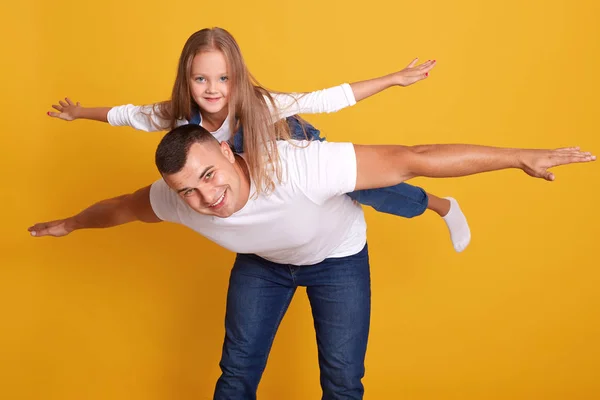 Ayah bahagia memberikan putrinya kuda-kudaan naik, ayah dan anak bersenang-senang bersama-sama, model berpose terislolasi atas latar belakang kuning di studio, keluarga bahagia memakai pakaian kasual. Konsep kebersamaan . — Stok Foto