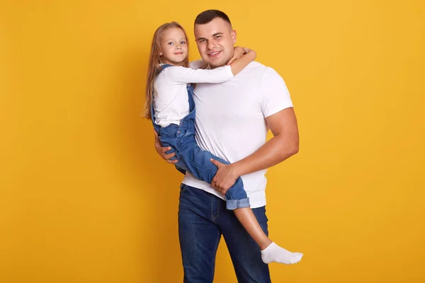 Στούντιο πλάνο του γοητευτικό κορίτσι παιδί με τον πατέρα της, όμορφος άντρας κρατώντας το παιδί στα χέρια, φορώντας casual ρούχα, θέτοντας απομονωμένη πάνω από κίτρινο φόντο Οικογένεια, τον τρόπο ζωής και την έννοια ευτυχία. — Φωτογραφία Αρχείου