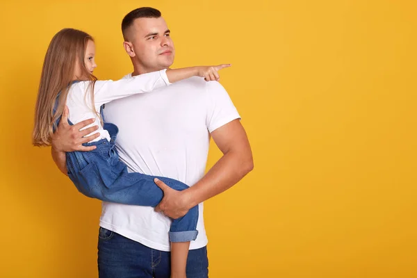 Profile view of young father hugging his little daughter and looking away, adorable female kid point aside with her finger, showing something to her daddy far away. Studio shot over yellow background. — Stock Photo, Image