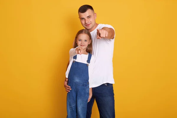 Horizontal shot of young attarctive man standing and smiling with his daughter, daddy and charming girl pointing in to camera, wearing casual clothing. Copy space for advertisment or promotion. — Stock Photo, Image