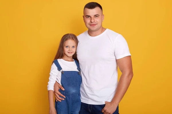 Bild av lycklig vit man omfamna sin charmiga lilla dotter, isolerade över gul studio bakgrund, pappa och hans barn poserar tillsammans, tittar direkt på kameran, bär casual kläder. — Stockfoto