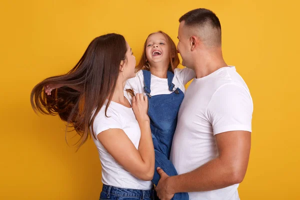 Imagen de una hermosa familia joven abrazándose y mirándose, riéndose mientras están de pie contra el fondo amarillo del estudio, divirtiéndose juntos, se ve feliz, expresa alegría. Concepto familiar . — Foto de Stock