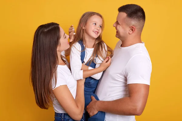 Indoor shot of happy young family with one child looking and smiling at camera, posing isolated over yellow studio background, spend time together, expressing happyness and love. Relationship concept.