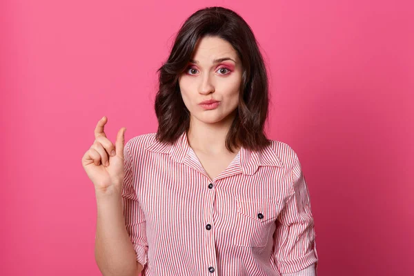 Halve lengte portret van vrouw tonen kleine hoeveelheid van iets en met niezende gezichtsuitdrukking, poseren geïsoleerd over rooskleurige achtergrond, goed uitziende dame dragen gestreept shirt. Mensenconcept. — Stockfoto