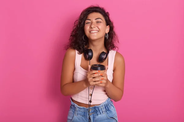 Ung vacker kvinna med hörlurar på halsen, ser glad, står leende isolerad över rosa studio bakgrund, attraktiv flicka bär rosa topp. Teknik, livsstil och människor koncept. — Stockfoto