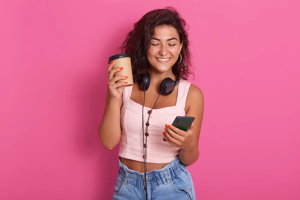 Chica alegre tomando un descanso de café, la lectura de la notificación en su teléfono móvil, actualizaciones aplicación favorita, escribiendo mensaje a su amigo y sonrisas, usando rosetón y jeans, aislado sobre fondo rosa . — Foto de Stock