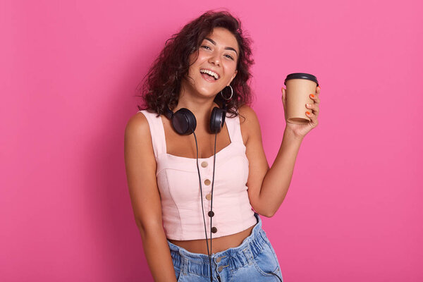 Close up portrait of cheerful young caucasian female with wavy dark hair, wearing rosy top and jeans, posing isolated on pink background. Adorable woman holds in hand paper cup with coffee and smiles.