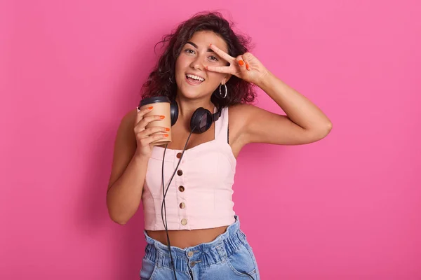 Foto di divertente donna caucasica veste top roseo e jeans, coprendo il suo occhio con segno di pace e tenendo in mano tazza di carta, vuole ascoltare musica con gli auricolari, isolato su sfondo rosa in studio . — Foto Stock
