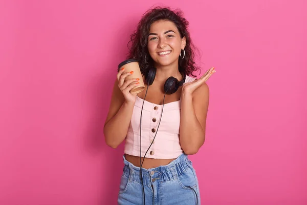 Horisontell bild av vacker kvinna poserar med toothy leende, hålla ta bort kaffekopp isolerad över rosa bakgrund, sprida en hand åt sidan, med mörkvågigt hår, klänningar topp och jeans. — Stockfoto