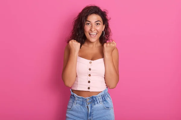 Foto de una joven feliz con un hermoso cabello oscuro y ondulado gritando y apretando el puño mientras mira directamente a la cámara, vistiendo un atuendo elegante, aislado sobre un fondo rosa. Gente emociones concepto . —  Fotos de Stock