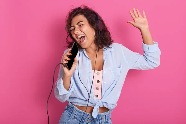 Close up portrait of young girl listening, enjoying music and holding cellular mp3 player in hand like microphone, wearing dance pink top, blue shirt and jeans, isolated over pink background. — Stock Photo, Image
