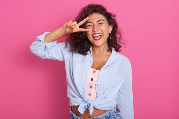 Cheerful woman showing peace sign with her fingers, beautiful young female with curly hair showing victory sign and looks smiling directly at camera isolated over pink background. Gesture concept/ — 스톡 사진