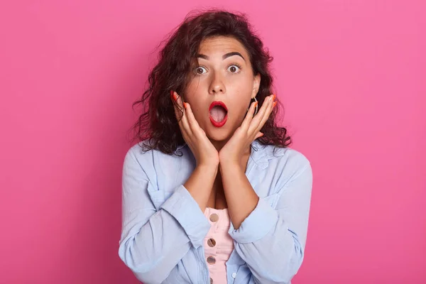 Imagen de la bonita mujer vistiendo camisa azul manteniendo las manos cerca de las mejillas, chica adorable posando con la boca ampliamente abierta, mira a la cámara con expresión de la cara asombrada mientras está de pie sobre el fondo rosado . — Foto de Stock