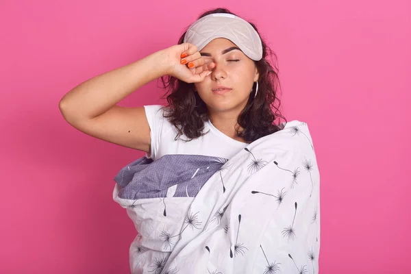 Porträtt av ung dam poserar med slutna ögon, har lust att sova, täcker sitt öga med handen, står under filt, bär sovmask, modell poserar isolerad över rosa vägg. Viloprincip. — Stockfoto