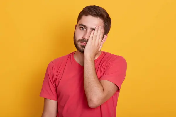 Das Bild eines gutaussehenden Mannes in lässigem rotem T-Shirt, der mit traurigem Gesichtsausdruck dasteht und sein halbes Gesicht mit einer Hand bedeckt, sieht müde aus, unrasiert und isoliert vor gelbem Hintergrund. Depressionskonzept. — Stockfoto