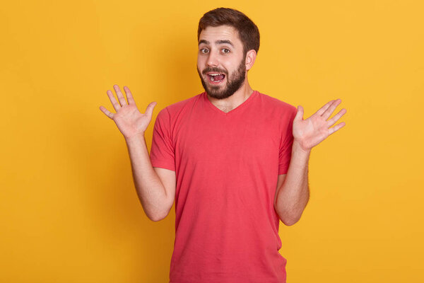 Portrait of man being surprised, handsome male spreading his hands up, posing isolated over yellow background, attractive unshaven guy wearing red casual t shirt. The concept of human emotions.