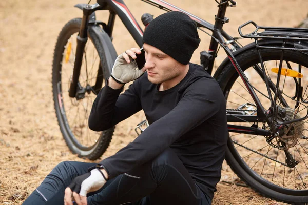 Close-up portret van fietser rusten in de open lucht, zitten op de grond in het veld of het bos en praten over de telefoon, draagt sportkleding en pet, het hebben van een serieus gesprek met vriend of vrouw tijdens het fietsen. — Stockfoto