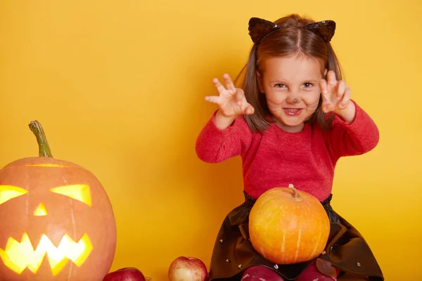 Imagen de una linda niña vestida con suéter rojo y falda negra, posando con orejas de gato y bigote, asustando a la gente frente a ella, sentada sobre un fondo amarillo del estudio. Halloween concepto de vacaciones . —  Fotos de Stock