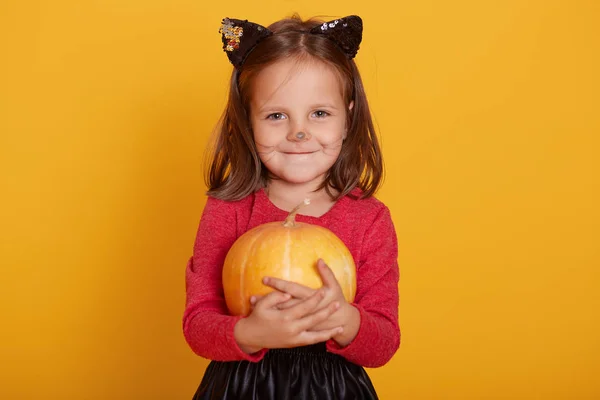 Retrato de niña vistiendo suéter rojo, con bigote y orejas de gato, niño encantador está sonriendo aislado sobre el fondo amarillo del estudio, sosteniendo calabaza naranja en las manos. Concepto Halloween . —  Fotos de Stock