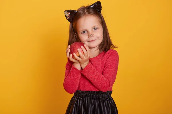 Retrato de cerca de una niña con traje de gato sosteniendo manzana envenenada roja, mirando directamente a la cámara, posando aislada sobre un fondo amarillo, con orejas de gato y bigote de gato pintado . —  Fotos de Stock