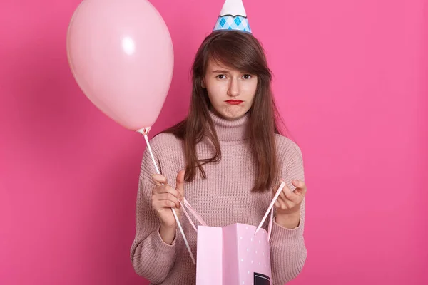 Retrato de close-up de bela menina decepcionada com pacote de presente e balão, vestindo suéter rosa e chapéu de aniversário, posando isolado sobre fundo rosado com lábios malvados, tendo expressão infeliz . — Fotografia de Stock