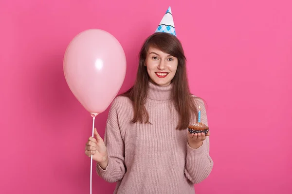 Горизонтальний знімок чудової дівчини з чарівною посмішкою, позує з капелюхом на день народження, тримає рожеву кульку і пиріг зі свічкою, виглядає щасливо, дивлячись прямо на камеру, ізольовано на рожевому. Концепція дня народження . — стокове фото