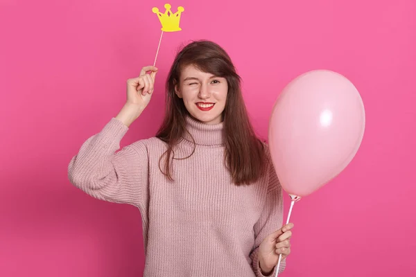 Portret van flirterige aantrekkelijke knipogende donkerharige vrouw met roze ballon, jurken warme roos trui, poseren tegen rooskleurige studio muur, hebben fotosessie en houden speelgoed kroon boven haar hoofd. — Stockfoto