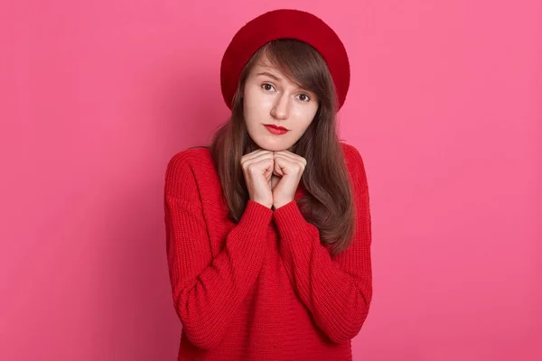 Image of caucasian darkhaired modest woman wearing red beret and sweater, model posing isolated over rosy background in studio, keeps fists under chin, looking at camera with sad expression.